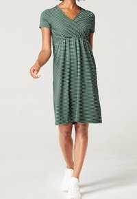 Grün gestreiftes Kleid mit einem V-Ausschnitt, kurzen Ärmeln und einer gerafften Taille. Hergestellt aus weichem Material, reicht es bis zur Mitte des Oberschenkels. Getragen mit weißen Sneakern.