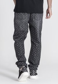 Jeans de mezclilla negra con un logotipo de patrón repetido en diferentes tonos, que cuentan con dos bolsillos traseros y piernas ajustadas, combinados con zapatillas negras.