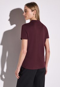 Burgundy, figursydd topp med kort ärm och rund halsringning. Slät tyg, minimala sömmar och rak fåll. Svarta byxor delvis synliga.