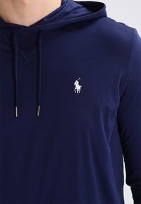 Námorní modrá mikina s kapucí vyrobená z měkké tkaniny, má klokaní kapsu, nastavitelné šňůrky a malé bílé logo na hrudi.