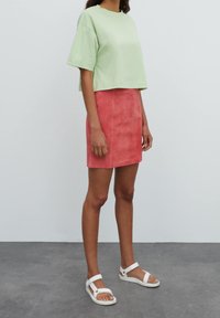 Camiseta corta de color verde claro con manga corta, combinada con una mini falda de ante rosa. Sandalias blancas de tiras completan el atuendo.