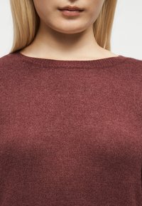 Gros plan d'une personne portant un pull en tricot bordeaux avec un col rond et des cheveux blonds visibles.