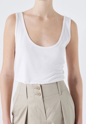 Femme portant un top blanc sans manches à col rond, rentré dans un pantalon beige plissé taille haute avec deux boutons.