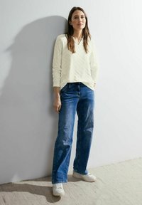 Helle Cremefarbene Sweatshirt mit einem strukturierten Zickzackmuster, kombiniert mit blauen Jeans und weißen Sneakers, vor einem neutralen Hintergrund.