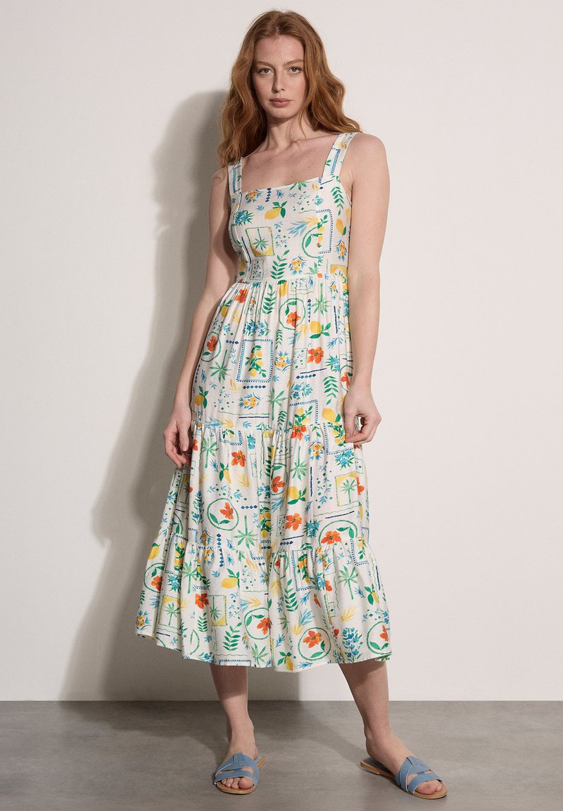 Femme debout contre un mur, portant une robe blanche sans manches à motif floral, de longueur moyenne, avec des sandales bleues à bout ouvert.
