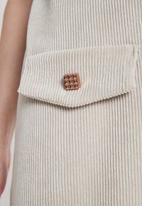 Beige Cordstoff mit vertikalen Rippungen, ausgestattet mit einer Klappentasche, die mit einem metallischen, strukturierten quadratischen Knopf verziert ist.