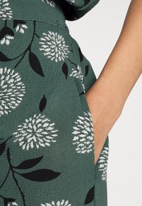 Tessuto verde e texture, con grandi motivi floreali bianchi e neri. Tasca laterale visibile, con una texture della pelle liscia sul bordo.