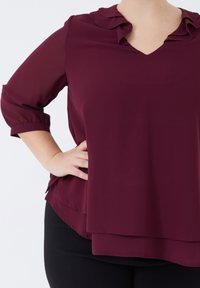 Bordeaux blouse met een V-hals en gerimpelde kraag, driekwart mouwen en gelaagd ontwerp; valt soepel met een gladde textuur.
