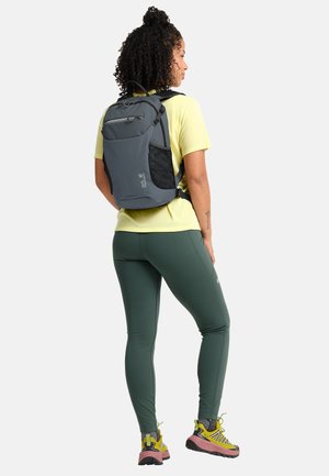 Donna che indossa una maglietta gialla, leggings verdi, scarpe da trekking gialle e zaino grigio, in piedi rivolta verso destra su sfondo bianco.