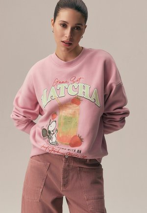 Žena nosící růžovou mikinu s grafikou tenisu s postavičkou Snoopyho a nápisem "Game, Set, Matcha", doplněnou tlumeně růžovými kalhotami.