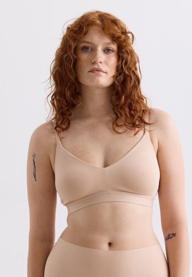 Frau mit roten lockigen Haaren und Sommersprossen, die einen hautfarbenen nahtlosen BH und hochgeschnittene Unterwäsche vor einem einfarbigen Hintergrund trägt.