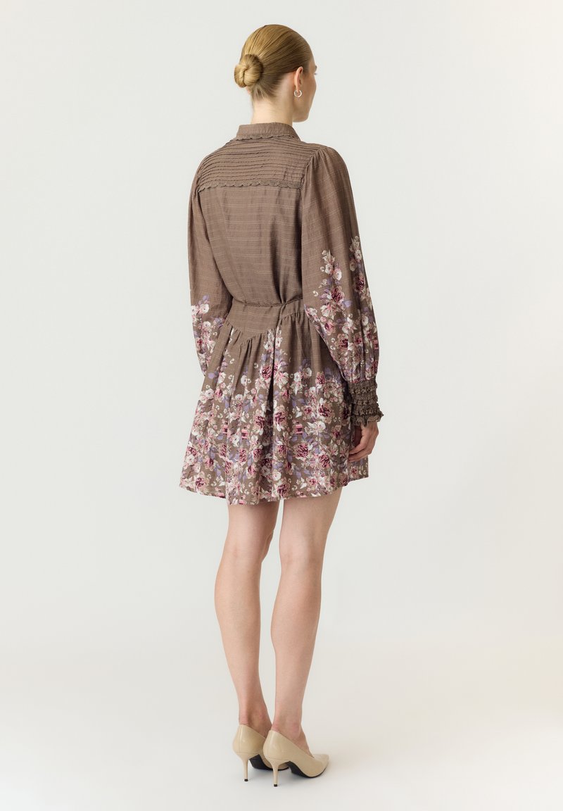 Femme avec un chignon blond portant une robe longue marron à fleurs à manches longues et des talons beiges, debout de dos sur un fond clair uni.