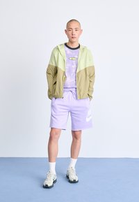 Mannenoutfit bestaande uit een lichtgroene en bruine windbreaker, een paars met patroon T-shirt, paarse shorts en witte sneakers met zwarte accenten.