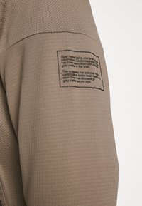 Bruine sportshirt met een mesh-textuur, voorzien van een geprinte label op de schouder met informatie over hersengezondheid en oefensuggesties.