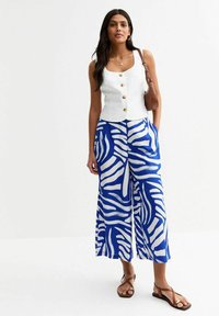 Top bianco con bottoni sul davanti e scollatura quadrata, abbinato a pantaloni larghi blu con un motivo astratto bianco e sandali piatti marroni.