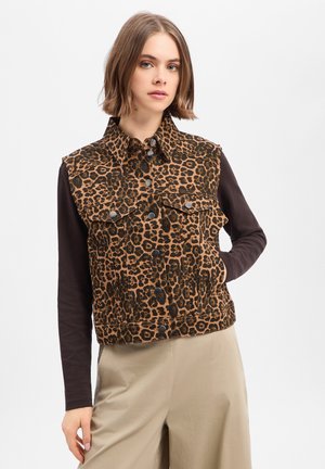 Jeune femme aux cheveux courts portant un gilet marron à imprimé léopard boutonné sur une chemise noire à manches longues et un pantalon beige, main dans la poche.