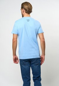 Svetlo modra bombažna majica s kratkimi rokavi, okroglim izrezom in majhnim logotipom na hrbtni strani. Nosi se z temno modrim džinsom.