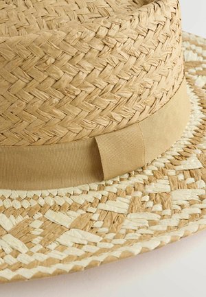 Široká klobúk z tkaného slamy v krémovej a svetlo hnedej farbe. Má hladký, béžový textilný pás okolo koruny.