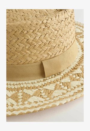 Široká klobúk z tkaného slamy v krémovej a svetlo hnedej farbe. Má hladký, béžový textilný pás okolo koruny.