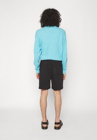 Turquoise textured sweater with long sleeves, paired with black shorts and sandals. Visible back textures and a patch pocket on the shorts.