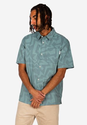 Man with braided hair wears teal short-sleeve button-up shirt with abstract patterns, beige pants, and looks downward with hands clasped.