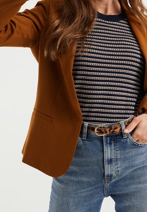 Femme portant un blazer marron, une chemise rayée bleu marine et blanche, un jean bleu et une ceinture à imprimé léopard avec boucle dorée, main reposant dans la poche.