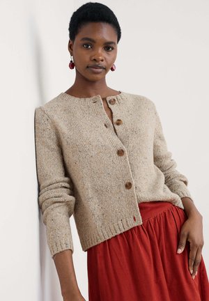 Young woman leaning against wall wearing beige knit buttoned cardigan, red skirt, and maroon geometric earrings, looking at camera.