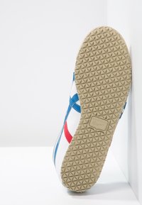 Semelle de basket blanche avec des bandes bleues et rouges, semelle extérieure en caoutchouc texturée beige, placée debout contre un mur et un sol blancs.