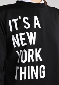 Sudadera negra con texto en bloque blanco que dice "ES UN ASUNTO DE NUEVA YORK". Cuenta con un escote redondo y una textura suave y lisa.