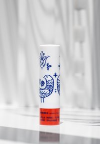 Vit cylindrisk läppbalsamrör med blå blommor och fågelmotiv. Röd bas med svart text som anger produktens namn och typ.