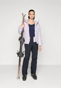 Γυναίκα σε σκινητό μπουφάν, παντελόνι και μπότες, κρατώντας τα σκι όρθια μπροστά σε λευκό φόντο.