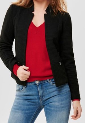 Kobieta ubrana w czerwony sweter z dekoltem w serek pod czarną kurtką w połączeniu z niebieskimi dżinsami, trzymająca kurtkę otwartą jedną ręką.