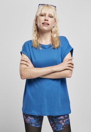T-shirt en coton bleu à manches courtes roulées, associé à des leggings à motifs avec des designs en pixels roses et bleus. La modèle se tient avec les bras croisés.