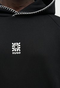 Μαύρο φούτερ με κουκούλα, με λευκό βελονιά βρόχου κατά μήκος της άκρης της κουκούλας και μικρό λευκό λογότυπο λουλουδιού με κείμενο "HUGO" στο στήθος.