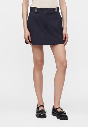 Femme portant une mini-jupe bleu marine à fines rayures avec des détails de boucle dorée et des chaussures de style Mary Jane noires avec des boucles.