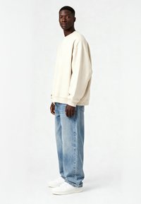 Bežinis per didelis džemperis su nuleistais pečių siūlais, derinamas su šviesiai mėlynomis laisvo kirpimo džinsais ir baltais sportbačiais. Lygi medžiaga, kasdieniškas dizainas.