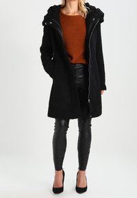 Musta keinoturkis takki hupulla, vetoketjulla ja pitkillä hihoilla, yhdistettynä mustiin keinonahkahousuihin ja mustiin korkokenkiin. Teksturoitu ruskea neule takin alla.