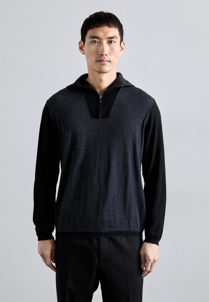 Férfi sötétszürke, zipzáras pulóverben fekete ujjakkal és fekete nadrágban áll egy sima világosszürke fal előtt.