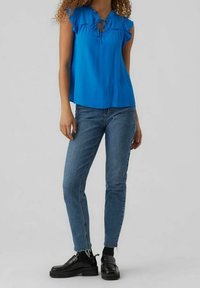 Donna che indossa una blusa blu senza maniche con rouches, jeans blu con orli sfrangiati, calzini neri e mocassini neri, in piedi su uno sfondo grigio.