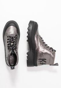 Botas de combate plateadas metálicas con suela de goma negra, punta redonda, acabado texturizado y cordones negros. Presenta un logo en el talón y una lengüeta.
