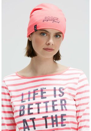 Pink cotton beanie with "SOC CX Original Brand" text. Model wears a striped pink and white long-sleeve shirt, featuring bold black lettering.