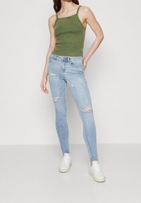 Top de tirantes finos ajustado verde combinado con jeans ajustados light blue con detalles desgastados y zapatillas blancas. Diseño minimalista.