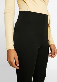 Höga svarta byxor gjorda av elastisk tyg, med en slät yta och skräddarsydd passform, matchade med en långärmad beige topp.