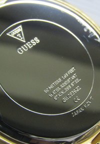 Fond de montre en acier inoxydable avec une surface noire polie. Le texte gravé inclut la marque "Guess", la classe de résistance à l'eau et les informations sur le modèle.