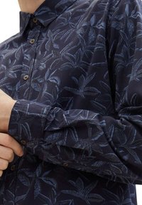 Hemd aus marineblauem Stoff mit blauem Blättermuster, mit Kragen, langen Ärmeln und braunen Knöpfen an der Vorderseite.