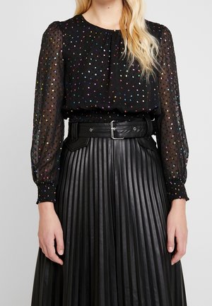 Donna che indossa una blusa nera trasparente a maniche lunghe con puntini multicolori e una gonna nera in pelle plissettata a vita alta con una cintura larga.