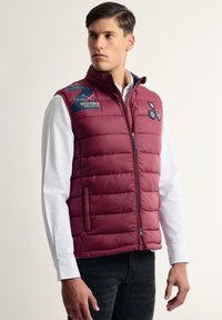 Chaleco acolchado burdeos con cierre de cremallera, bolsillos laterales y detalle del logo en el hombro izquierdo; presenta un diseño acolchado y una textura suave.
