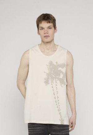 Ung mann med lys hud og kort hår iført en ermeløs beige skjorte med en dempet palmetregrafikk, stående foran en ensfarget bakgrunn.