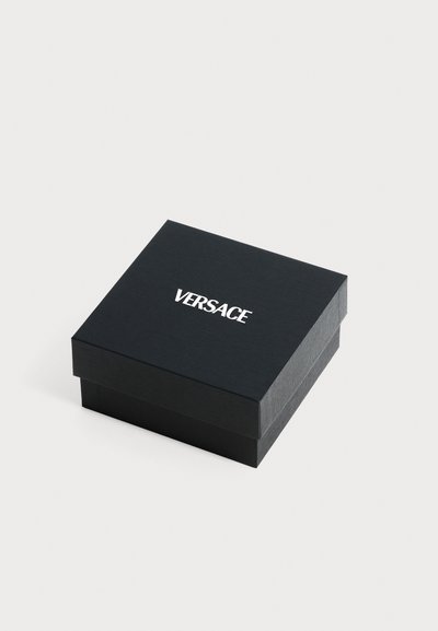 Boîte carrée noire de marque Versace avec un logo blanc centré sur le couvercle, posée sur un fond uni gris clair.