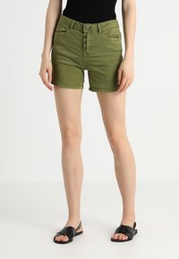 Pantalones cortos de mezclilla verde con un diseño ajustado, cinco bolsillos y cierre de botón. Textura suave con un aspecto ligeramente desgastado. Sandalias negras tipo slide.